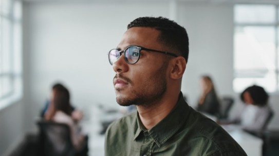 Close-up of a young African American male employee wearing eyeglasses contemplating in office about how to stand out in a competitive job market.