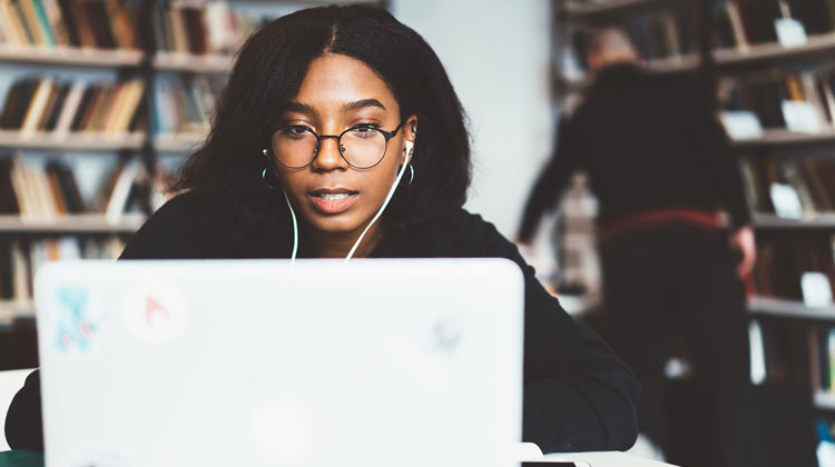 In the school library, a young African American female high school student wearing vision protection eyewear works on a laptop computer, concentrating, and completing the college application process.
