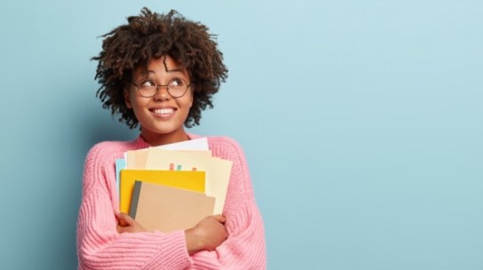 Your African-American female student with natural hair is holding folders and notebooks in front of her. She has glasses on and is wearing a pink sweater.