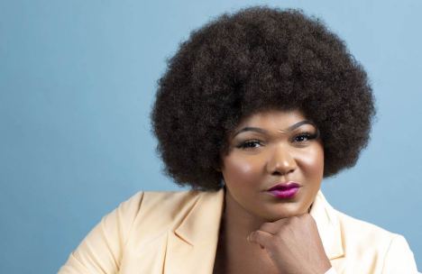 Confident academic professional, Dr. Jessica Love, with an afro, posing thoughtfully in a cream blazer against a light blue background.